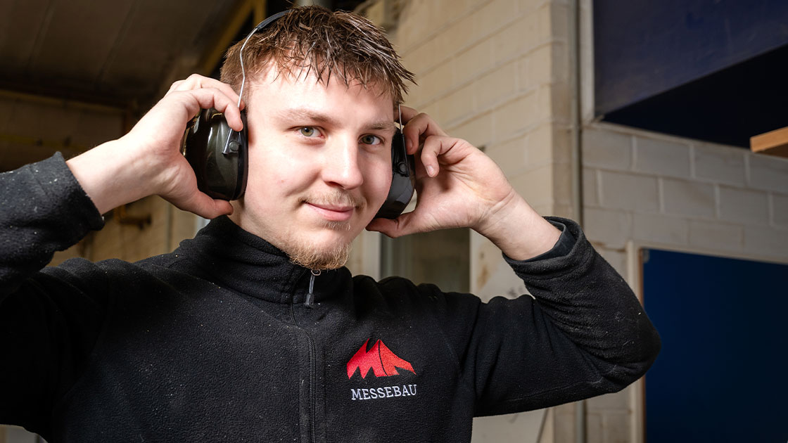 MA Messebau Mitarbeiter Fertigung mit den Händen am Gehörschutz